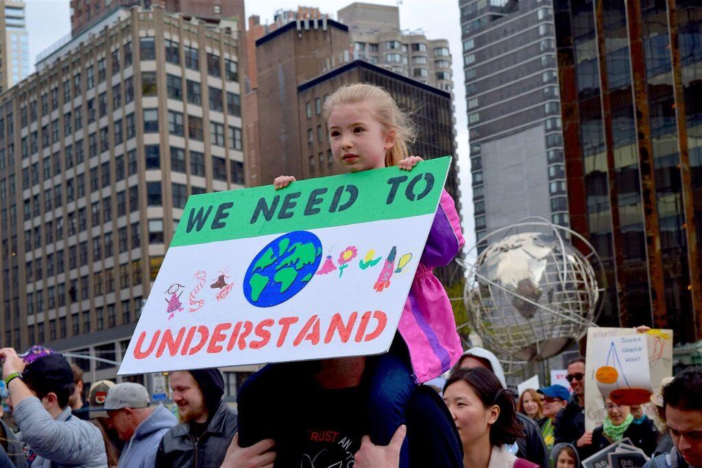 Petite fille en manifestation portant une banderole "We need to understand". Traduction : Nous avons besoin de comprendre.